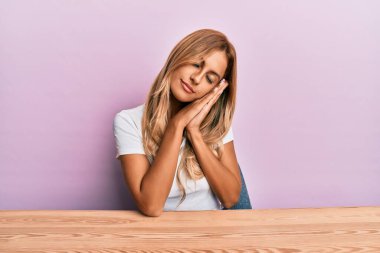 Güzel sarışın genç kadın gündelik giysiler içinde masada oturuyor yorgun uyuyor hayal kuruyor ve elleri ile poz verirken kapalı gözlerle gülümsüyor.. 