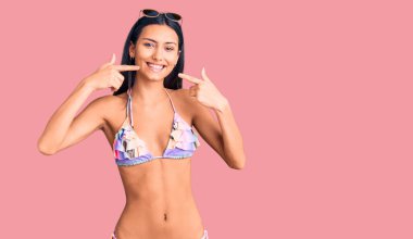 Bikini giyen, güneş gözlüklü neşeli gülümseyen, parmaklarıyla ve ağzıyla işaret eden güzel Latin bir kız. diş sağlığı kavramı. 
