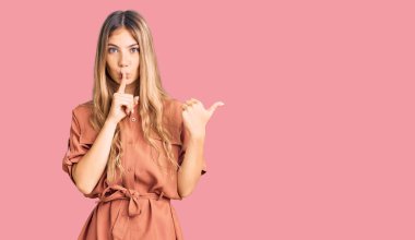 Sarı saçlı, yaz tulumu giyen güzel, beyaz bir kadın parmakları dudaklarında, elleri yandan gösteren bir şekilde sessiz olmak istiyor. sessizlik ve gizli konsept. 