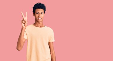 Genç Afro-Amerikalı günlük kıyafetler giymiş iki numarayı gösterip işaret ederken kendinden emin ve mutlu gülümsüyor.. 