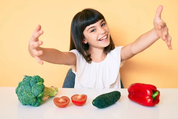 年轻的小女孩 带着砰的一声坐在桌子上 蔬菜一样地看着镜头 张开双臂拥抱着 欢快的表达拥抱幸福 — 图库照片