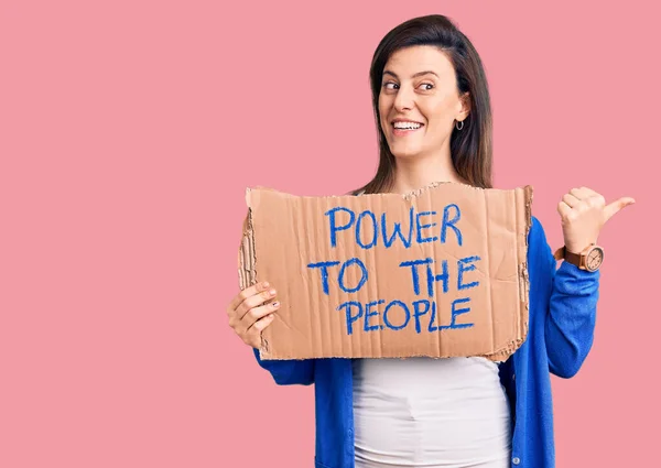 Young beautiful woman holding power to the people banner pointing thumb up to the side smiling happy with open mouth