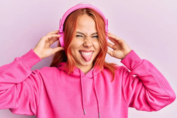 用耳机听音乐的红头发年轻女人带着滑稽的表情高兴地伸出舌头 — 图库照片