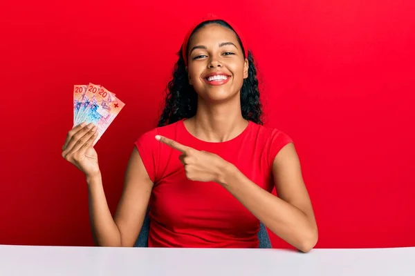 年轻的非洲裔美国姑娘手握瑞士法郎钞票坐在桌子上 高兴地微笑着 手指手画脚 — 图库照片