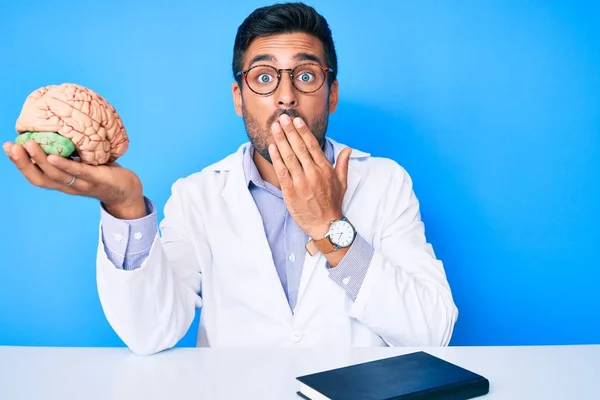 Jovem Hispânico Sentado Mesa Usando Casaco Médico Segurando Cérebro Cobrindo — Fotografia de Stock