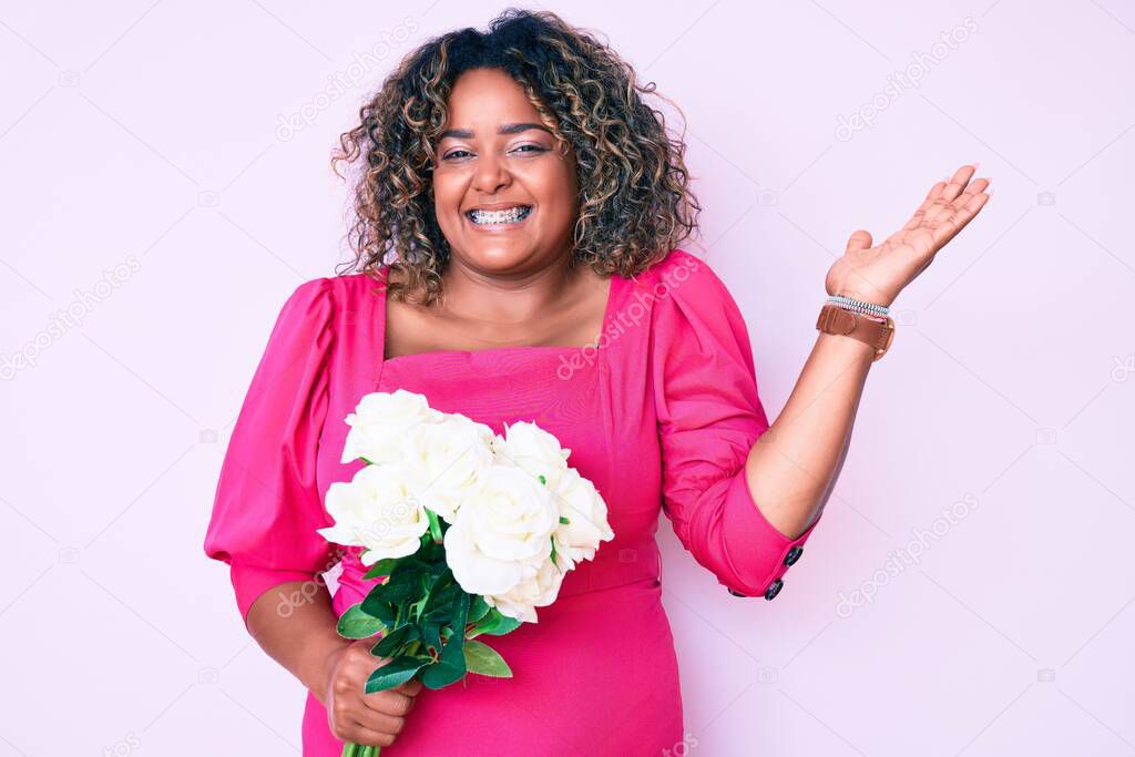 Young african american plus size woman holding flowers celebrating victory with happy smile and winner expression with raised hands 