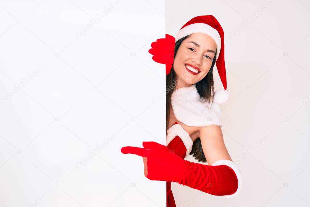Young woman wearing santa claus costume holding blank empty banner smiling happy pointing with hand and finger 