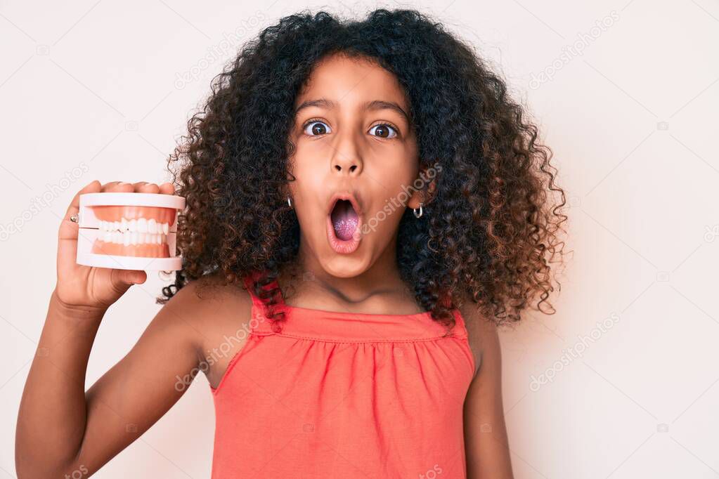 African american child with curly hair holding denture scared and amazed with open mouth for surprise, disbelief face 