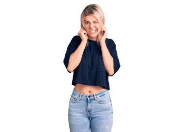 Gürültülü müzik sesi için kulaklarını parmaklarıyla kapatan gündelik tişört giyen genç, güzel sarışın kadın. sağır kavramı. 