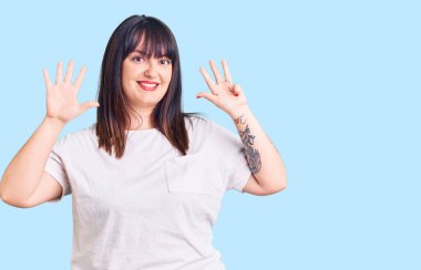 Genç ve cüsseli kadın gündelik kıyafetler giyip dokuz numaralı parmaklarıyla işaret ederken kendinden emin ve mutlu bir şekilde gülümsüyor.. 