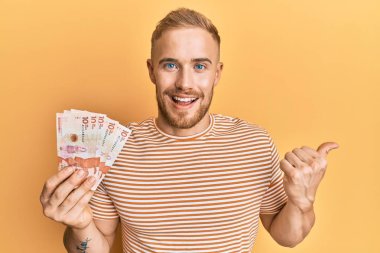 Elinde 10 Kolombiya pezosu olan beyaz bir adam başparmağıyla yan tarafa bakıyor ve ağzı açık gülümsüyor. 