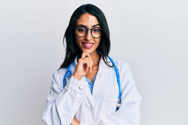 Doktor üniforması ve steteskop giymiş güzel İspanyol bir kadın kameraya kollarını kavuşturup çenesini kapatarak gülümsüyor. Olumlu düşünmek. 