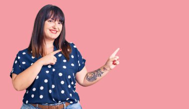 Gündelik kıyafetler giyen, gülümseyen ve kameraya bakan, iki eli ve parmağı yanda olan genç bir kadın.. 