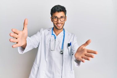 Doktor üniforması ve steteskop giymiş genç yakışıklı adam kameraya sarılıp gülümsüyor. Mutluluğu kucaklayan neşeli bir ifade.. 