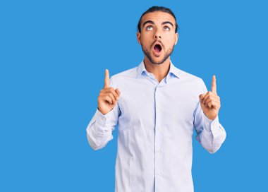 İş kıyafetleri giymiş genç yakışıklı adam şaşırmış ve şaşırmış bir şekilde yukarı bakıyor ve parmaklarıyla işaret ediyor ve kollarını kaldırıyor.. 