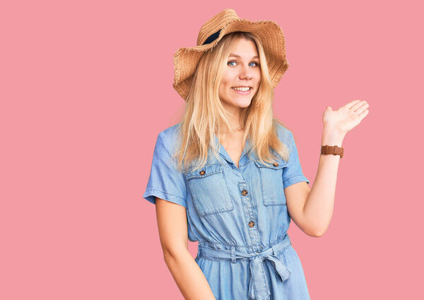 Молодая красивая блондинка в летней шляпе и платье, улыбаясь веселым подарком и указывая ладонью на камеру. 