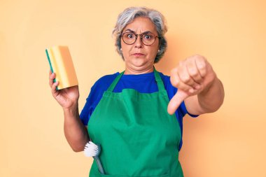 Yaşlı Latin asıllı, gri saçlı, önlük takan kızgın yüzlü, olumsuz işaretli, baş parmakları aşağıda, reddedilme konsepti olan bir kadın. 