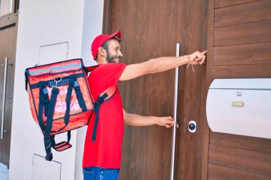 Beyaz teslimatçı, kırmızı üniforma giyiyor ve sırt çantası teslim ederken gülümsüyor.