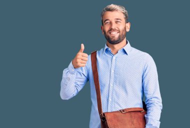 Zarif bir gömlek giyen sarışın genç adam evrak çantası tutuyor mutlu ve pozitif gülümsüyor, başparmağıyla mükemmel ve onay işareti yapıyor. 