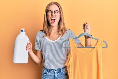 Askıda tişört tutan güzel sarışın kadın ve deterjan şişesi kızgın ve kızgın çığlık atan sinirli ve öfkeli, öfkeyle bağıran.. 