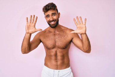 Genç İspanyol adam tişörtsüz duruyor ve parmaklarıyla on numarayı gösterirken kendinden emin ve mutlu bir şekilde gülümsüyor.. 