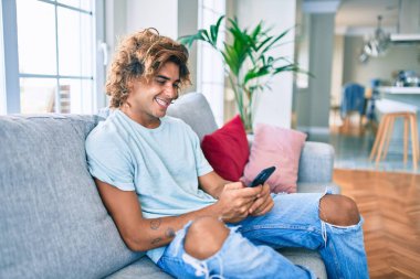 Genç İspanyol adam evde akıllı telefon kullanarak mutlu bir şekilde gülümsüyor.