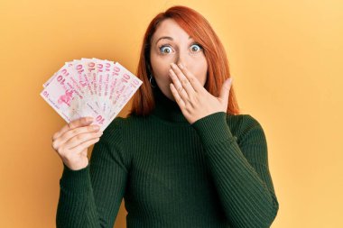 Güzel kızıl saçlı kadın elinde 10 lira banknotla ağzını eliyle kapatıyor, şaşırmış ve hata yapmaktan korkuyor. şaşırmış ifade 