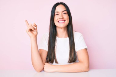 Masada oturan beyaz tişörtlü genç beyaz kadın gülümsüyor mutlu bir şekilde elini ve parmağını yana doğru gösteriyor. 