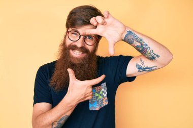 Yakışıklı, kızıl saçlı, uzun sakallı, sıradan kıyafetler ve gözlüklü, gülümseyen, elleri ve parmakları mutlu yüzlü bir adam. yaratıcılık ve fotoğrafçılık kavramı. 