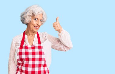 Önlük takan yaşlı, gri saçlı, mutlu ve pozitif gülümseyen, başparmağıyla mükemmel ve onay işareti yapan bir kadın. 