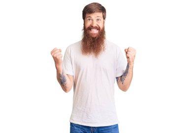 Uzun sakallı, beyaz tişörtlü yakışıklı genç adam kolları açık ve kolları açık bir şekilde başarıyı kutluyor ve şaşırıyor. Kazanan konsept. 