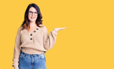 Güzel, genç, esmer, günlük kıyafetler ve gözlük takan neşeli bir şekilde gülümseyen ve avuç içi kameraya bakan bir kadın.. 