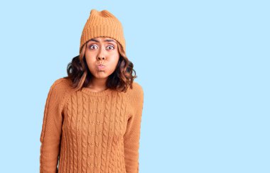 Genç, güzel, melez, yün kazak giyen, şapkalı, komik yüzlü bir kadın. Havayla şişmiş ağız, çılgın bir ifade.. 