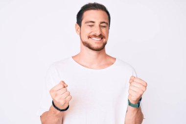 Başarılı olmak için heyecanlı, kolları havada ve gözleri kapalı zaferi kutlayan günlük beyaz tişört giyen genç yakışıklı bir adam. Kazanan konsept. 