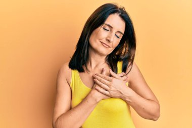 Orta yaşlı esmer kadın, sarı arka plan üzerine günlük kıyafetler giyip gülümsüyor, elleri göğsünde, gözleri kapalı, yüzünde minnet dolu bir jestle. sağlık kavramı. 