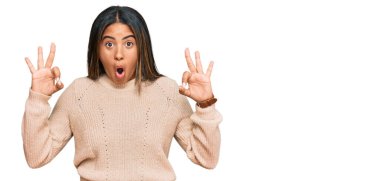 Kışın yün kazak giyen genç Latin kız şaşırmış ve şaşırmış görünüyor. Tamam sembolünü parmaklarıyla yapıyor. çılgın ifade 