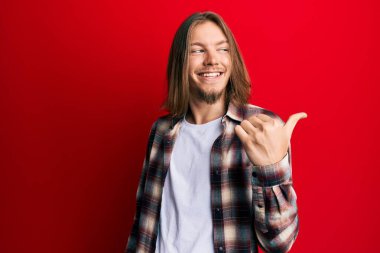 Uzun saçlı yakışıklı, sıradan kıyafetler giyen, gülen yüzlü gülümseyen ve baş parmağıyla yüzünü gösteren beyaz bir adam.. 