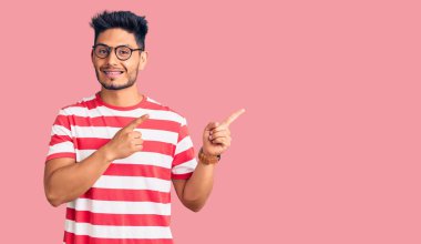 Yakışıklı Latin Amerikalı genç adam günlük kıyafetler ve gözlük takıyor gülümsüyor ve kameraya bakıyor iki eliyle ve parmaklarıyla yandan işaret ediyor.. 