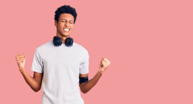 Genç Afrikalı Amerikalı adam spor kıyafetleri giyiyor ve kulaklık takıyor. Çok mutlu ve heyecanlı. Kollarını kaldırıp, gülümseyerek ve başarı için bağırarak kazanan jestler yapıyor. kutlama konsepti. 