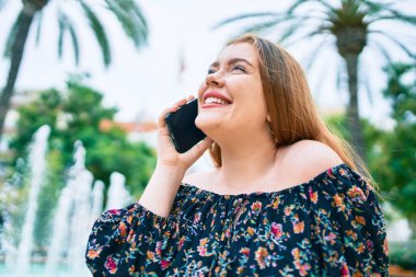 Genç kızıl saçlı kadın parkta akıllı telefondan konuşuyor.