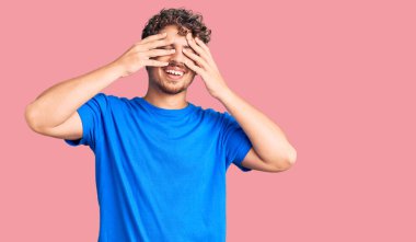 Kıvırcık saçlı, sıradan kıyafetler giyen, gözleri kapayan neşeli ve komik gülümseyen genç yakışıklı bir adam. kör kavram. 