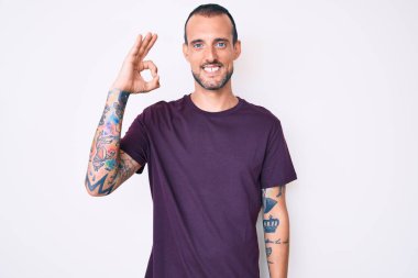 Dövmeli, sıradan kıyafetler giyen, olumlu gülümseyen, el ve parmaklarıyla imza atan genç yakışıklı bir adam. başarılı ifade. 