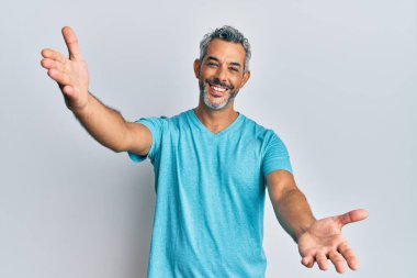 Orta yaşlı, gri saçlı, sıradan kıyafetler giyen, kameraya bakıp sarılmak için kollarını açmış gülümseyen bir adam. Mutluluğu kucaklayan neşeli bir ifade.. 