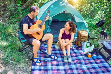 Ormanda kamp yaparken gülümseyen orta yaşlı bir çift. Yerde oturup klasik gitar çalarak şarkı söylemek.