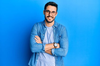 Gündelik kıyafetler ve gözlük takan genç İspanyol adam kameraya bakarken kollarını kavuşturup gülümsüyor. pozitif kişi. 