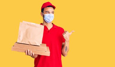 Genç, yakışıklı, İspanyol asıllı, yemek dağıtan güvenlik maskesi takan, başparmağını yana doğru gösteren, ağzı açık gülümseyen bir adam. 