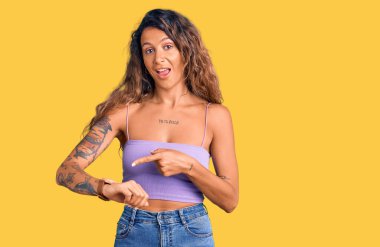 Dövmeli genç İspanyol kadın. Günlük kıyafetler giyiyor. Aceleyle saati, sabırsızlığı ve gecikmeyi gösteriyor. 