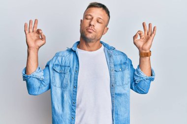 Sıradan kot ceket giyen yakışıklı kaslı adam rahat ol ve gözün kapalı gülümse parmaklarınla meditasyon hareketi yap. yoga konsepti. 