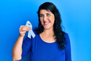 Artı mavi kurdeleli esmer kadın. Pozitif ve mutlu görünüyor. Kendine güvenen bir gülümsemeyle dişlerini gösteriyor. 