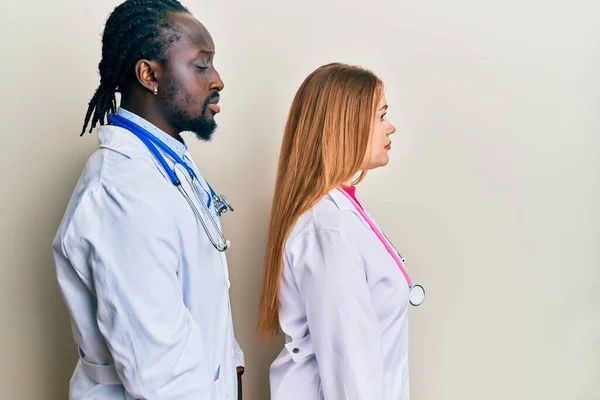 Young interracial couple wearing doctor uniform and stethoscope looking to side, relax profile pose with natural face with confident smile.